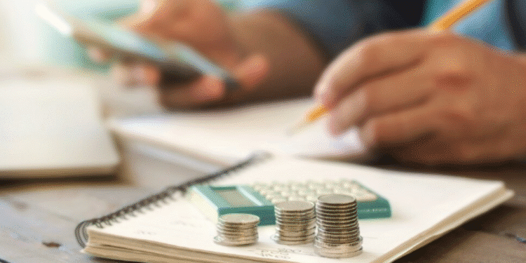 Pilha de moedas sobre caderno com calculadora ao fundo, enquanto pessoa usa celular e faz anotações.