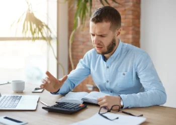 Homem com barba e camisa azul, sentado em uma mesa de escritório, com expressão de frustração, olhando para uma calculadora e um livro.