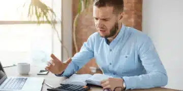 Homem com barba e camisa azul, sentado em uma mesa de escritório, com expressão de frustração, olhando para uma calculadora e um livro.