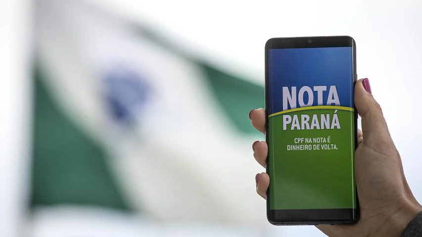 Pessoa segurando um celular com a tela exibindo a logo do Nota Paraná, com a bandeira do Brasil ao fundo