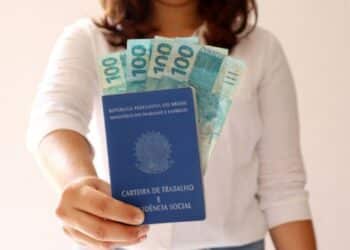 Mulher segurando a carteira de trabalho brasileira com notas de 100 reais visíveis.