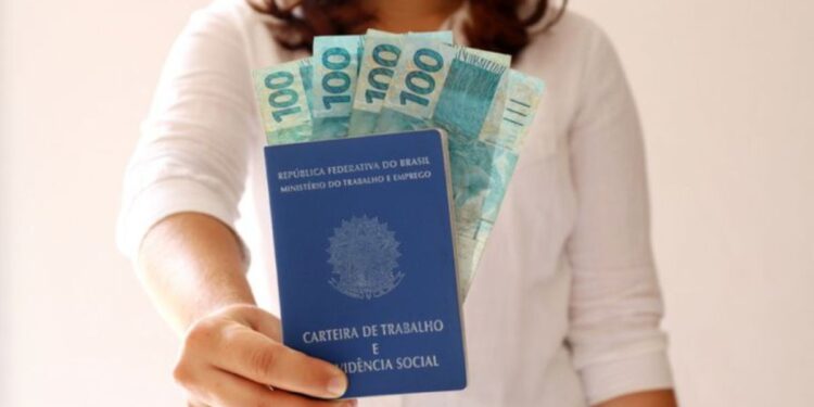 Mulher segurando a carteira de trabalho brasileira com notas de 100 reais visíveis.