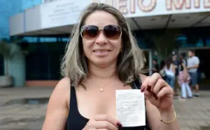 Mulher sorridente segurando um cupom fiscal, em frente a um edifício, comemorando um possível prêmio.