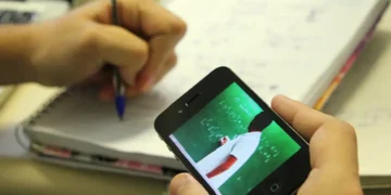 Pessoa assistindo aula online no celular enquanto faz anotações em caderno.