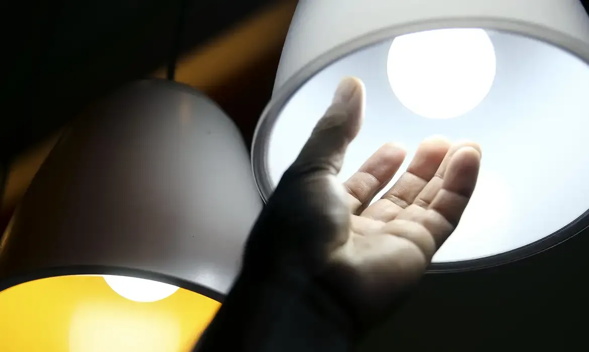 Mão estendida próxima a uma lâmpada acesa simbolizando a análise da conta de luz.