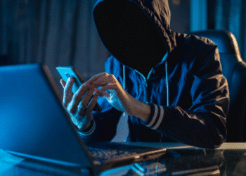 Pessoa encapuzada manipulando celular e laptop representando golpista digital