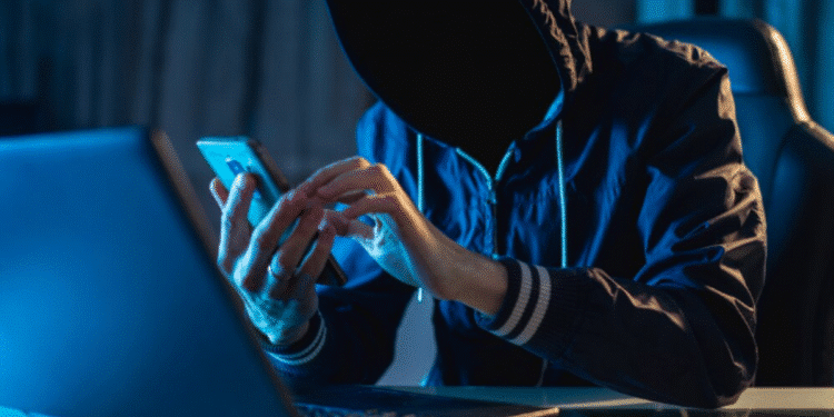 Pessoa encapuzada manipulando celular e laptop representando golpista digital