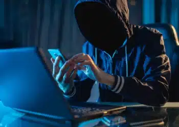 Pessoa encapuzada e sem rosto segurando celular e usando notebook, representando golpes virtuais e fraudes online em 2025.