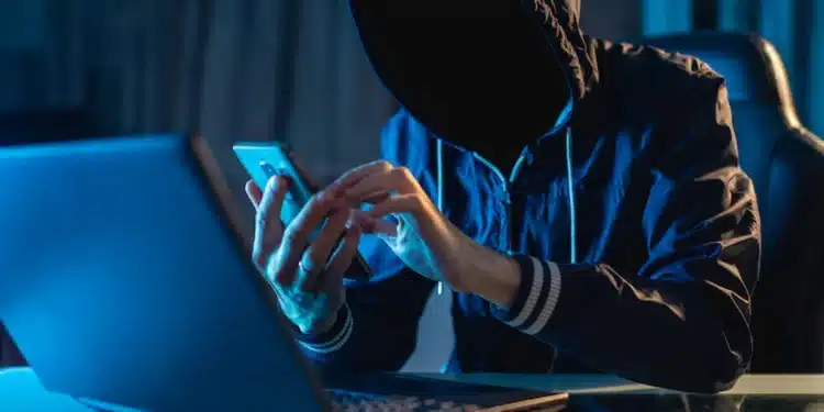 Pessoa encapuzada e sem rosto segurando celular e usando notebook, representando golpes virtuais e fraudes online em 2025.