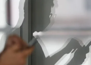 Imagem de uma sombra de uma pessoa refletindo na parede, mostrando as mãos mexendo no celular.