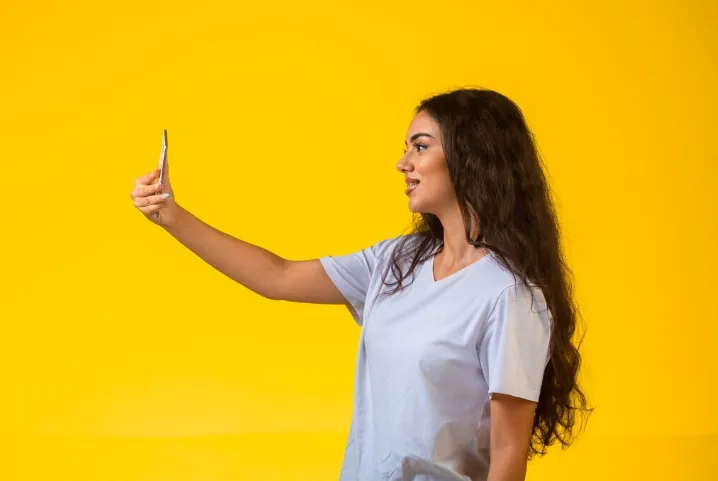 Mulher tirando selfie segurando o celular com fundo amarelo