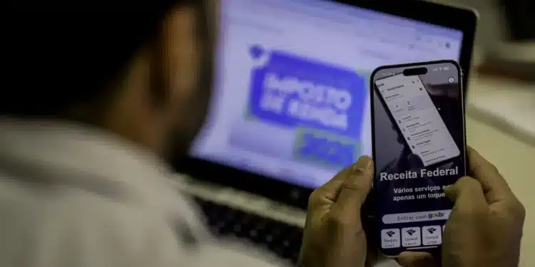 "Pessoa utilizando o aplicativo da Receita Federal no celular enquanto visualiza o site no computador, ilustrando a importância do envio dentro do prazo."