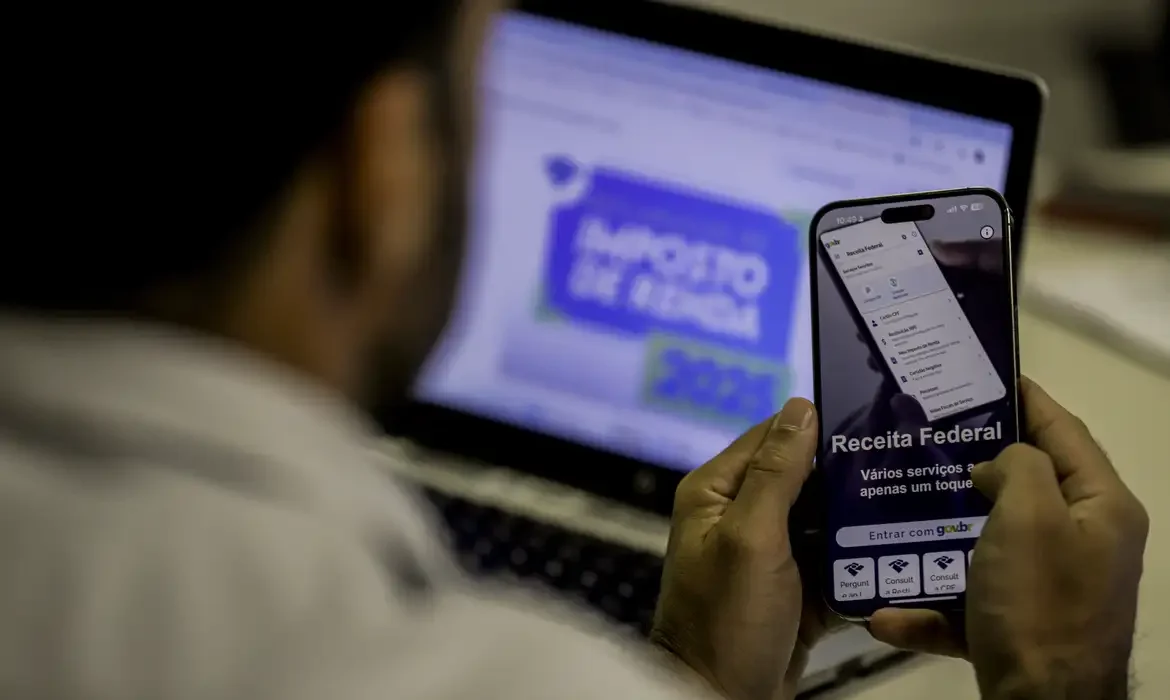 "Pessoa utilizando o aplicativo da Receita Federal no celular enquanto visualiza o site no computador, ilustrando a importância do envio dentro do prazo."