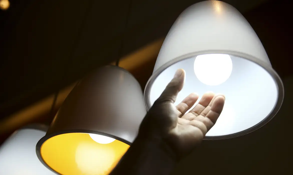 Mão se aproximando de uma luminária pendente com lâmpada acesa.