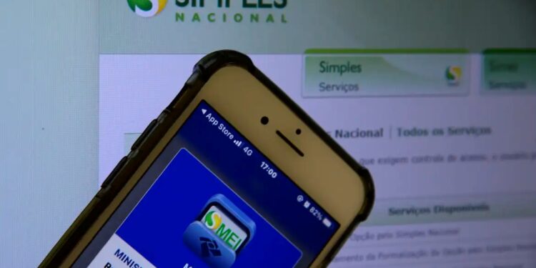 Imagem de um celular exibindo o aplicativo MEI da Receita Federal, com o site do Simples Nacional ao fundo, destacando a importância da declaração anual para MEI.