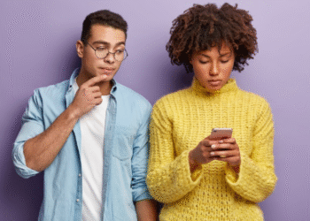 Jovem mulher concentrada usando celular, enquanto homem ao lado observa com expressão pensativa.