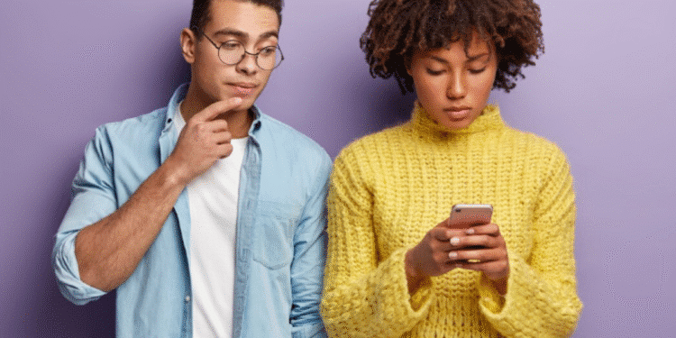 Jovem mulher concentrada usando celular, enquanto homem ao lado observa com expressão pensativa.