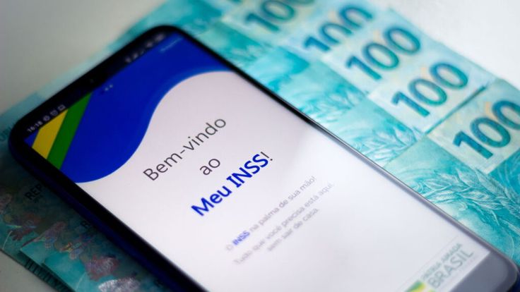 Aplicativo Meu INSS aberto em um celular sobre notas de 100 reais