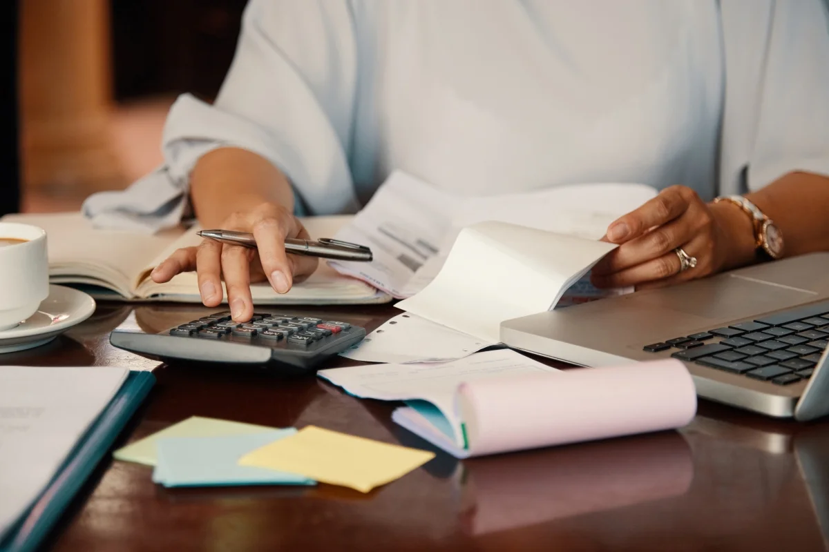 Pessoa trabalhando com calculadora e documentos em uma mesa de escritório, lidando com finanças pessoais.
