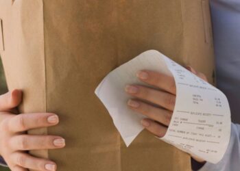 Pessoa segurando uma sacola de papel com compras e uma nota fiscal visível na mão.