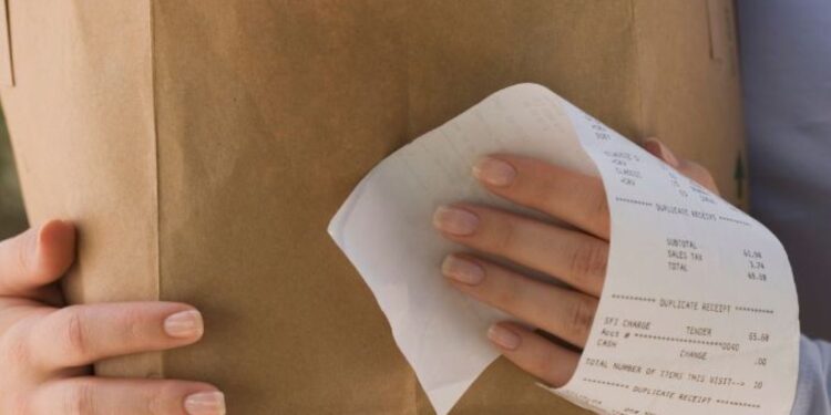 Pessoa segurando uma sacola de papel com compras e uma nota fiscal visível na mão.