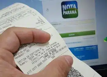 Imagem de um recibo do programa Nota Paraná sendo segurado em frente a uma tela de computador com a página de login