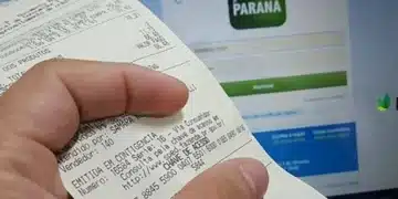 Imagem de um recibo do programa Nota Paraná sendo segurado em frente a uma tela de computador com a página de login