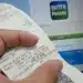 Imagem de um recibo do programa Nota Paraná sendo segurado em frente a uma tela de computador com a página de login