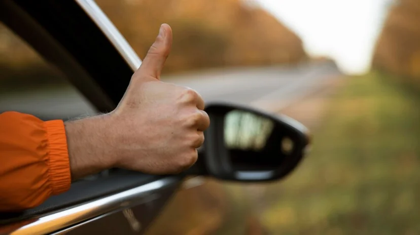 Pessoa fazendo sinal de positivo pela janela do carro, simbolizando aprovação ou conclusão bem-sucedida.