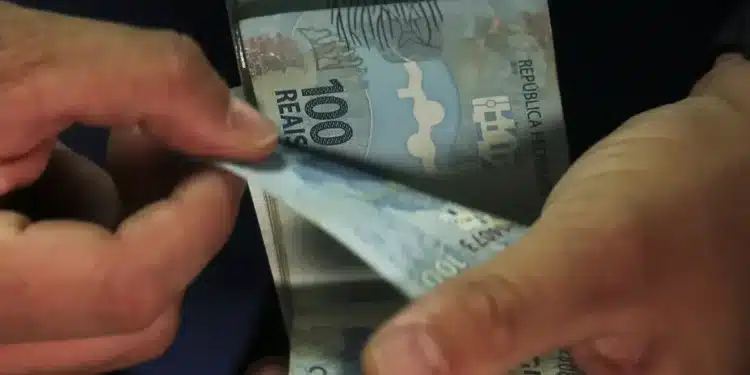 Mãos contando cédulas de 100 reais, ilustrando empréstimo consignado e portabilidade bancária.