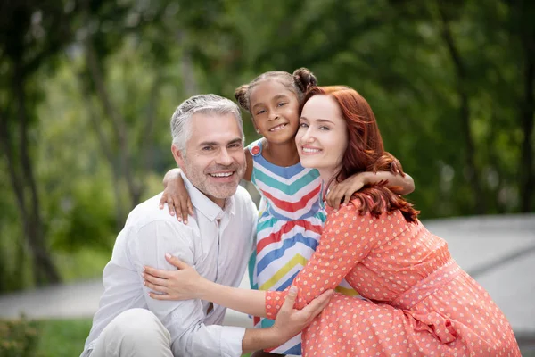 Família adotiva feliz com pais e criança abraçados em ambiente ao ar livre.