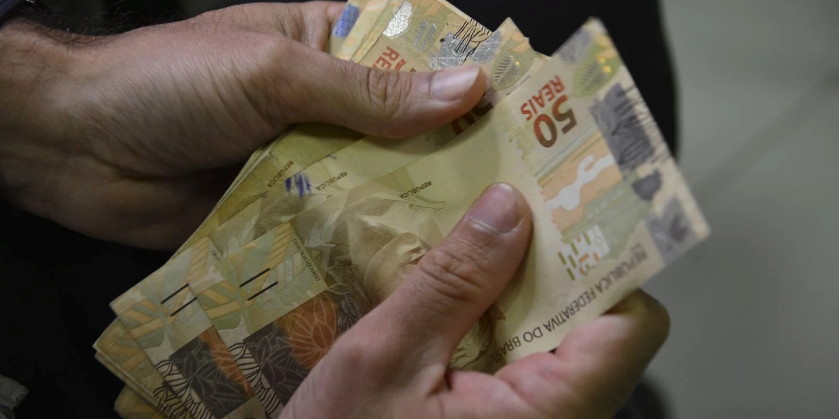Mãos segurando cédulas de R$ 50, representando a disparidade entre o salário mínimo e o valor necessário para uma vida digna.