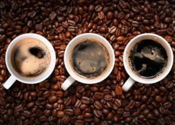 Três xícaras de café expresso vistas de cima, cercadas por grãos de café, com diferentes tons de crema na superfície.