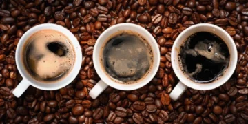 Três xícaras de café expresso vistas de cima, cercadas por grãos de café, com diferentes tons de crema na superfície.