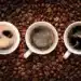 Três xícaras de café expresso vistas de cima, cercadas por grãos de café, com diferentes tons de crema na superfície.