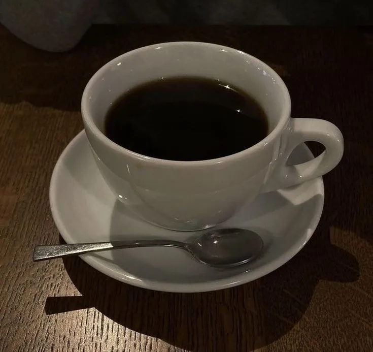 Xícara branca com café preto, acompanhada de colher de metal, sobre mesa de madeira escura.