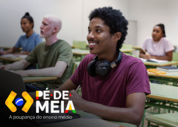 Estudante sorrindo enquanto usa um laptop em sala de aula, com outros alunos ao fundo, representando a temática do benefício educacional do Pé-de-Meia.