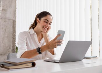 Mulher com celular e laptop sorrindo, simbolizando o trabalho remoto e online.