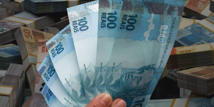 Mão segurando notas de 100 reais com pilhas de dinheiro ao fundo