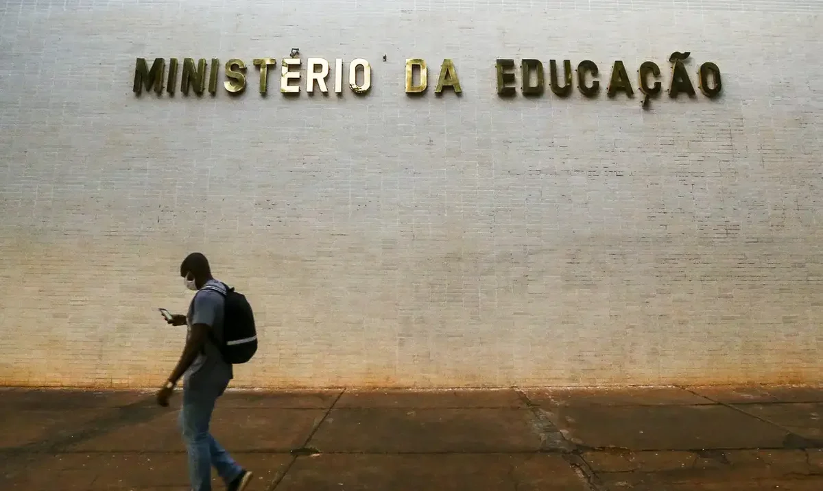 Pessoa caminhando em frente ao Ministério da Educação com celular, representando estudantes se beneficiando de auxílio educacional.