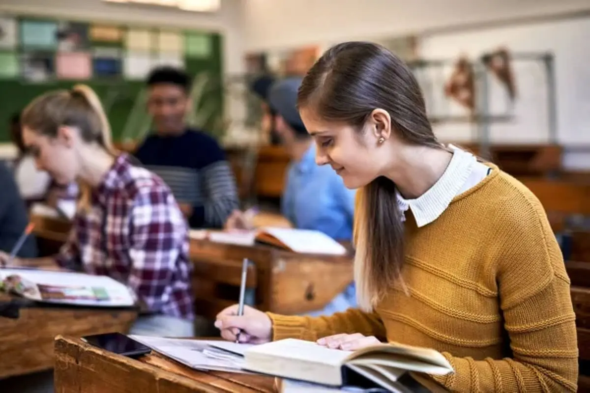 Estudante em sala de aula, anotando em seu caderno enquanto outros colegas de classe também estudam.