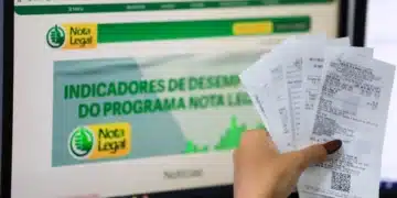 Mão segurando várias notas fiscais em frente à tela com o site do programa Nota Legal