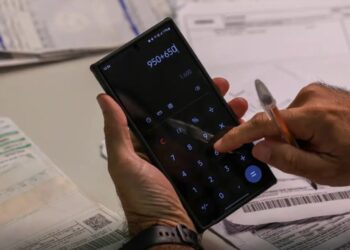 Pessoa usando calculadora no celular para somar valores de contas bancárias e boletos acumulados.