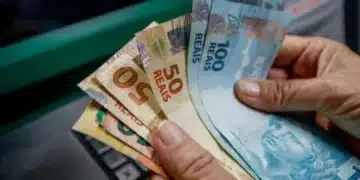 Dinheiro em reais sendo segurado por uma mão próxima a um caixa eletrônico.