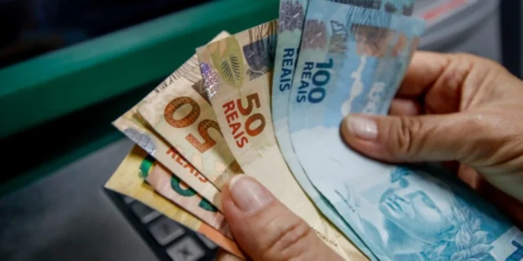 Dinheiro em reais sendo segurado por uma mão próxima a um caixa eletrônico.