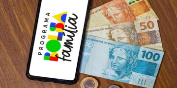 Aplicativo do Bolsa Família aberto no celular ao lado de cédulas e moedas brasileiras sobre mesa de madeira
