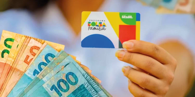 Cartão do Bolsa Família em mãos, com cédulas de reais ao fundo, representando o pagamento do programa social.