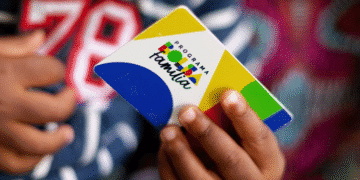 Cartão do Programa Bolsa Família sendo segurado por uma mão, com fundo colorido e texto sobre revisão de cadastro.