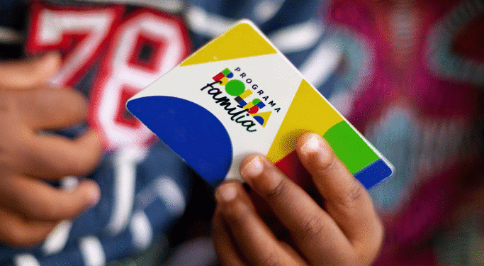 Cartão do Programa Bolsa Família sendo segurado por uma mão, com fundo colorido e texto sobre revisão de cadastro.