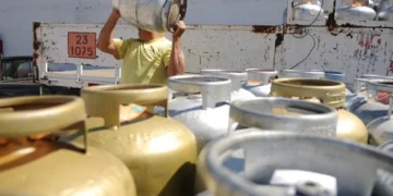 Homem carregando botijão de gás em uma área com várias unidades ao fundo.